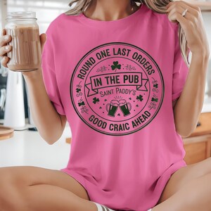 Azalea pink unisex t-shirt featuring the "Good Craic Ahead" pub seal design in dark green. A fun, playful twist on traditional St. Patrick's Day gear, perfect for women or men who want a softer color palette while keeping the Irish drinking humor.