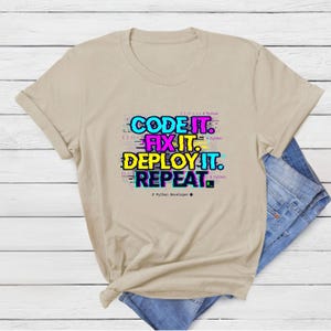 Sand beige tee with a colorful vibrant glitch-style programming quote. "Code Fix Deploy Repeat" graduation gift for Class of 2026 computer science majors.