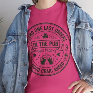 Heliconia pink unisex t-shirt featuring the "Good Craic Ahead" pub seal design in dark green. A fun, playful twist on traditional St. Patrick's Day gear, perfect for women or men who want a softer color palette while keeping the Irish drinking humor.