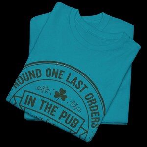 May include: Two folded teal t-shirts. The top shirt has a circular graphic with the text "ROUND ONE LAST ORDERS IN THE PUB" in dark green. A shamrock is in the center of the graphic.