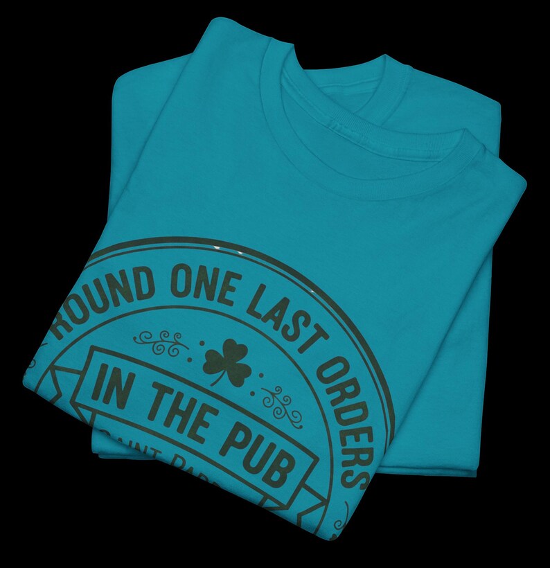 May include: Two folded teal t-shirts. The top shirt has a circular graphic with the text "ROUND ONE LAST ORDERS IN THE PUB" in dark green. A shamrock is in the center of the graphic.