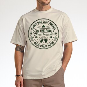 Sand beige unisex t-shirt with a dark green "Last Orders In The Pub" emblem. The vintage-style beer mug graphic and shamrock details give it a retro, worn-in Irish bar feel, ideal for a casual, understated St. Patrick's Day outfit.