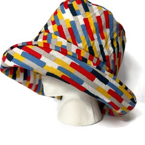 May include: A wide-brimmed hat with a colorful geometric pattern in shades of red, yellow, blue, and gray. The hat is made of a soft, textured fabric.