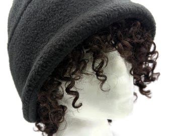May include: A dark gray fleece hat with a short brim, displayed on a white mannequin head. The hat has a textured surface and a rounded crown. Dark brown curly hair is visible beneath the hat, creating a contrast in color and texture.