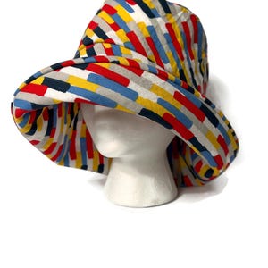 May include: A wide-brimmed bucket hat with a colorful geometric pattern. The hat is made of a lightweight fabric and features a variety of colors, including red, yellow, blue, and gray.