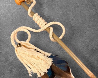 May include: A long, slender wooden wand featuring a smooth, light-brown finish. The top is adorned with a polished wooden sphere, a small metallic charm, and a thick, cream-coloured twisted rope that wraps around the shaft, ending in a tassel.