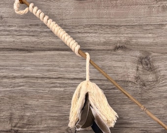 May include: A cat toy wand featuring a long, thin wooden stick with a small brown bead at the top. A thick, cream-colored twisted rope is wrapped around the upper portion of the stick, ending in a dangling tassel made of cream fringe and feathers.