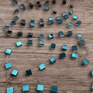 May include: A strand of square-shaped beads featuring a metallic finish in shades of teal, blue, and dark grey. The beads are strung on a thin, clear filament, creating a repeating pattern of reflective, geometric shapes along the length of the cord.