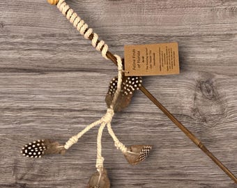 May include: A cat teaser toy featuring a long brown wooden wand with a cream-colored rope wrapped around the handle and a light wood bead at the end. Three cream rope strands hang from the wand, each tipped with a brown and white spotted feather.