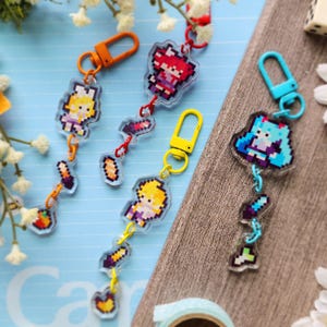 May include: A selection of pixel art keyrings depicting video game characters. Each keyring has a different coloured clasp and chain. The characters are rendered in a pixelated style, with details such as hair, clothing, and accessories. The keyrings are made of clear acrylic.