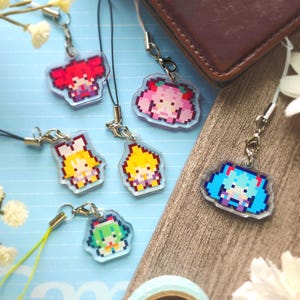 May include: A selection of pixel art acrylic charms depicting anime-style characters in various colours. Each charm has a silver clasp and a black or yellow lanyard. The charms are displayed on a light blue surface with a brown leather wallet in the background.