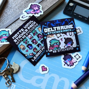 May include: Two sticker packs featuring characters from the video game Deltarune. The first pack is blue and white and says "Matte Vinyl Sticker Pack with 19 Stickers". The second pack is black and blue and says "DELTARUNE STICKER PACK" and "19 STICKERS".