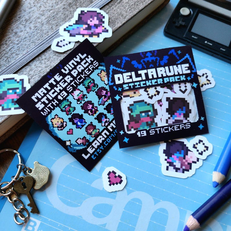 May include: Two sticker packs featuring characters from the video game Deltarune. The first pack is blue and white and says "Matte Vinyl Sticker Pack with 19 Stickers". The second pack is black and blue and says "DELTARUNE STICKER PACK" and "19 STICKERS".
