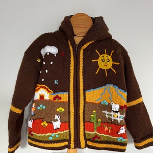 May include: Brown knitted hooded sweater with colourful embroidered details of a sun, clouds, a house, cacti, and llamas. The sweater has a zip closure and yellow and brown stripes on the sleeves and bottom.