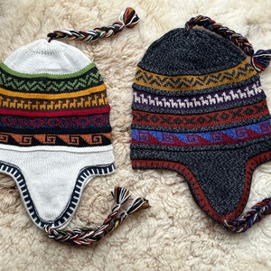 May include: Two knitted winter hats with ear flaps and braided tassels. One hat is white with colorful geometric patterns, the other is gray with similar patterns.