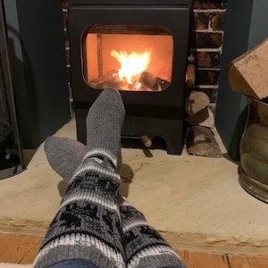 May include: A pair of gray and black patterned socks with white stripes are worn by a person sitting in front of a fireplace with a fire burning inside. The person's feet are resting on a stone hearth.