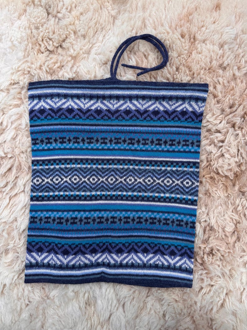 May include: A blue knitted tote bag with a geometric pattern in shades of blue, white, and black. The bag has a dark blue handle and is made of a soft, textured material. The bag is rectangular in shape.