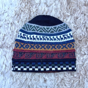 May include: A knitted beanie hat with a geometric pattern in navy blue, white, blue, green, gold, and burgundy. The hat features a repeating design of zigzags, diamonds, and squares. The hat is made of a soft, warm material.