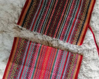 May include: Two rectangular, woven textile bags with vibrant, geometric patterns. The bags feature a mix of red, orange, black, gray, and tan stripes and diamond shapes. Each bag has a red strap attached. The bags are displayed on a fluffy, white surface.