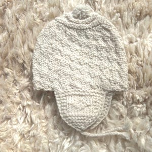 May include: Cream-colored knitted hat with ear flaps and a pom-pom on top. The hat features a textured diamond pattern and is made of a soft, warm material. The ear flaps extend down to provide extra warmth and coverage.