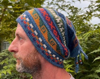May include: A blue knit beanie featuring a multi-colored geometric pattern in shades of orange, green, and white. The hat has a slouchy fit with dangling knit ties at the back, each finished with a small dark brown wooden bead at the end.