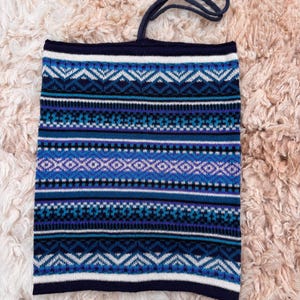 May include: A blue and white patterned neck gaiter with a drawstring. The gaiter features a geometric design in shades of blue, white, and purple. The background is a soft, light brown, textured surface.