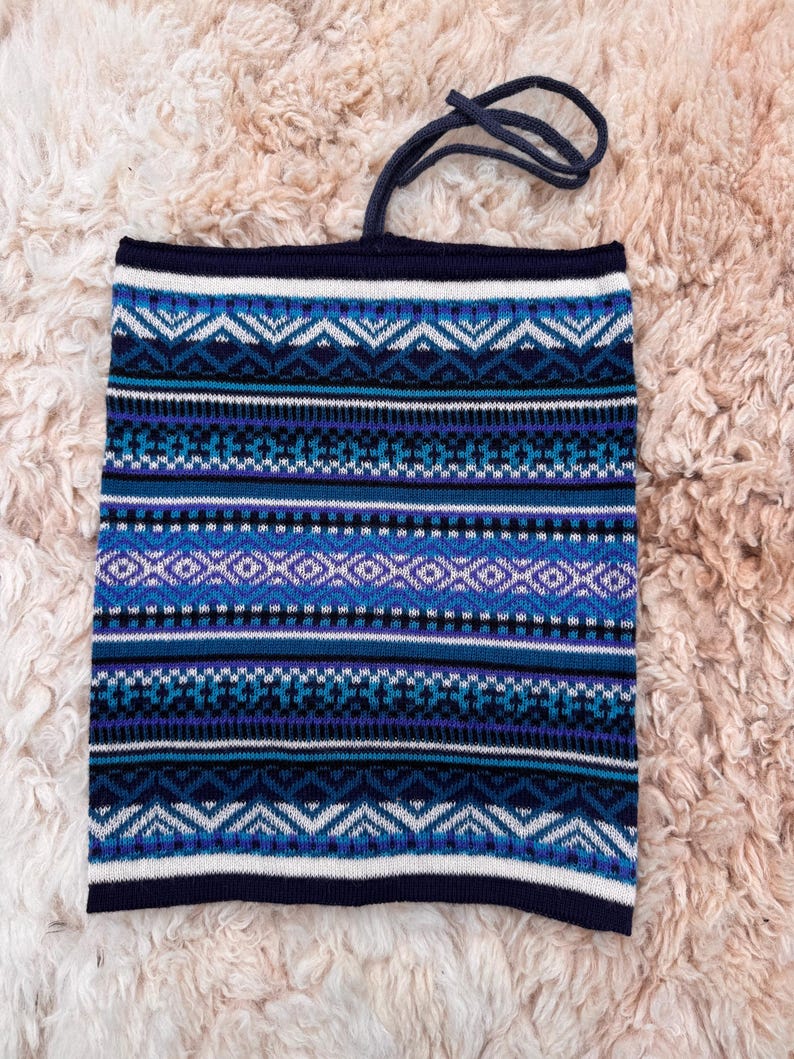 May include: A blue and white patterned neck gaiter with a drawstring. The gaiter features a geometric design in shades of blue, white, and purple. The background is a soft, light brown, textured surface.