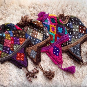 May include: Four brightly coloured, handmade crochet hats with ear flaps and tassels. Each hat features a patchwork design with squares of various colours, including pink, purple, blue, and brown. The hats are displayed on a fluffy, cream-coloured surface.