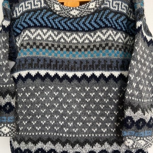 K&ouml;nnte beinhalten: Ein grauer, blauer und wei&szlig;er Strickpullover mit geometrischem Muster. Der Pullover hat einen Rundhalsausschnitt und lange &Auml;rmel.