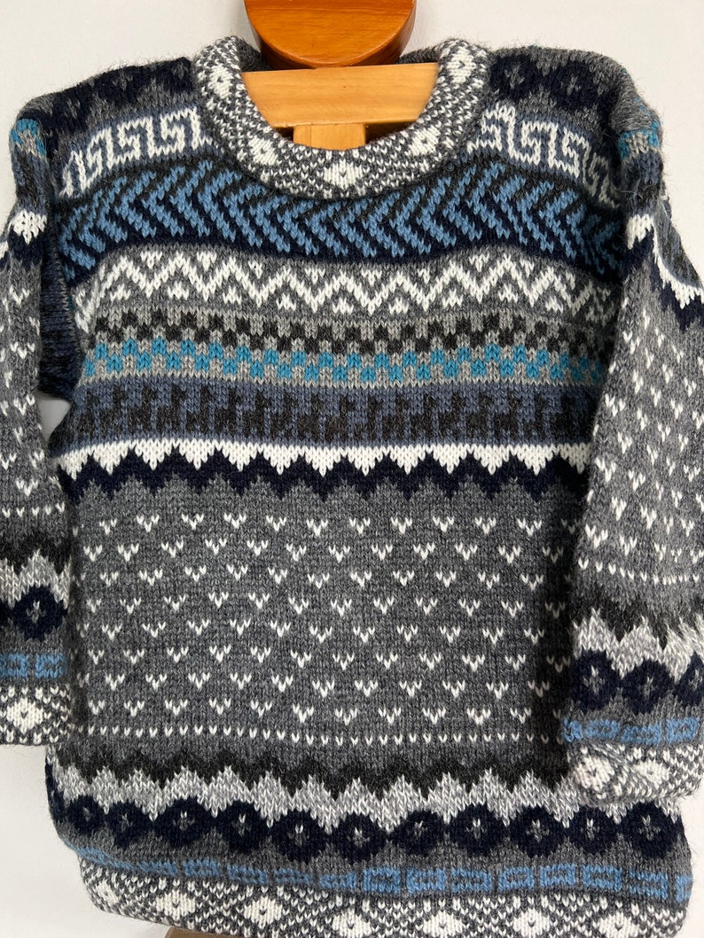 K&ouml;nnte beinhalten: Ein grauer, blauer und wei&szlig;er Strickpullover mit geometrischem Muster. Der Pullover hat einen Rundhalsausschnitt und lange &Auml;rmel.