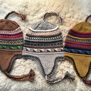 May include: Three knitted winter hats with ear flaps and long braids. The hats are brown, grey, and mustard yellow with colourful geometric patterns.