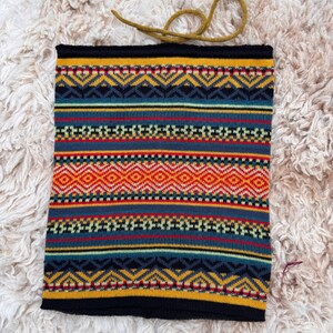 May include: A knitted neck gaiter with a geometric pattern in shades of yellow, blue, red, and orange. The neck warmer has a black border and a mustard-colored drawstring. The item is displayed on a fluffy, off-white surface.