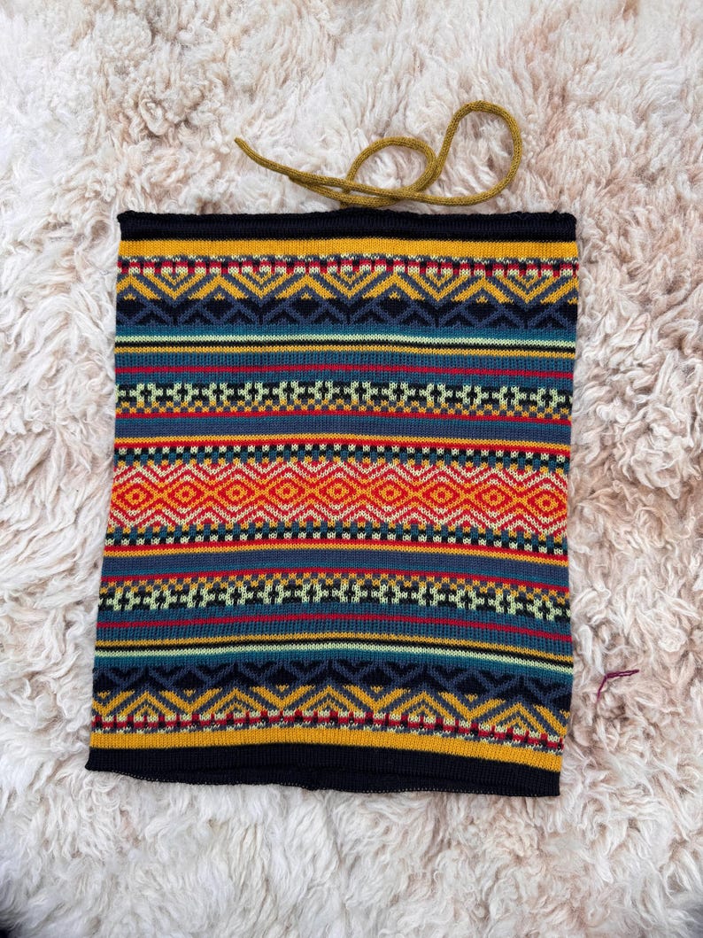 May include: A knitted neck gaiter with a geometric pattern in shades of yellow, blue, red, and orange. The neck warmer has a black border and a mustard-colored drawstring. The item is displayed on a fluffy, off-white surface.