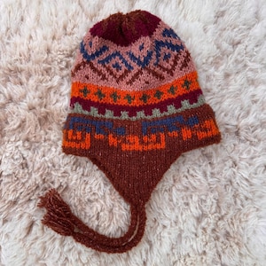 May include: A colorful knit hat with ear flaps and tassels. The hat features a geometric pattern in shades of orange, blue, brown, and pink. The hat is made of a soft, textured material, perfect for cold weather.