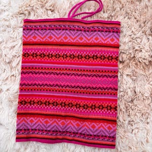May include: A vibrant pink and red knitted neck warmer with a geometric pattern. The design features horizontal bands of diamonds, zigzags, and stripes in shades of pink, red, and black. A pink cord is attached at the top.