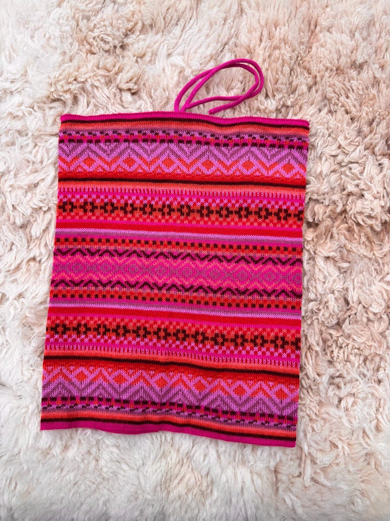 May include: A vibrant pink and red knitted neck warmer with a geometric pattern. The design features horizontal bands of diamonds, zigzags, and stripes in shades of pink, red, and black. A pink cord is attached at the top.
