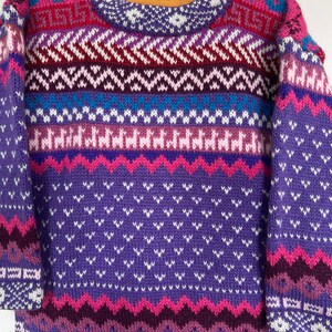 K&ouml;nnte beinhalten: Ein lila, rosa und blauer Strickpullover mit geometrischem Muster. Der Pullover hat einen Rundhalsausschnitt und lange &Auml;rmel.