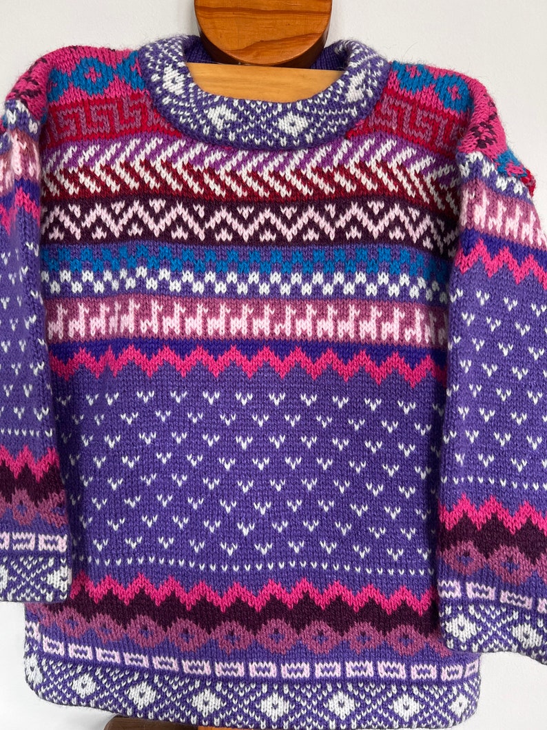 K&ouml;nnte beinhalten: Ein lila, rosa und blauer Strickpullover mit geometrischem Muster. Der Pullover hat einen Rundhalsausschnitt und lange &Auml;rmel.