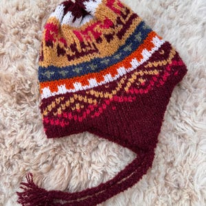 May include: A colorful knit hat with ear flaps and tassels. The hat features a pattern of geometric shapes in shades of burgundy, white, orange, blue, and gold. The tassels are burgundy. The hat is likely handmade and suitable for cold weather.