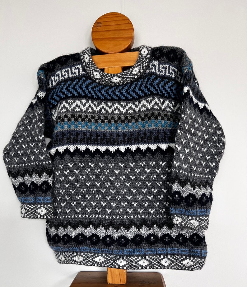 K&ouml;nnte beinhalten: Ein grauer, blauer und wei&szlig;er Pullover mit geometrischem Muster. Der Pullover hat ein geometrisches Muster mit einem sich wiederholenden Muster aus Dreiecken und Rauten.