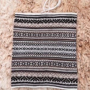 May include: A knitted neck gaiter with a geometric pattern in shades of brown, gray, and white. The gaiter has a white drawstring at the top. The background is a textured, light brown surface.
