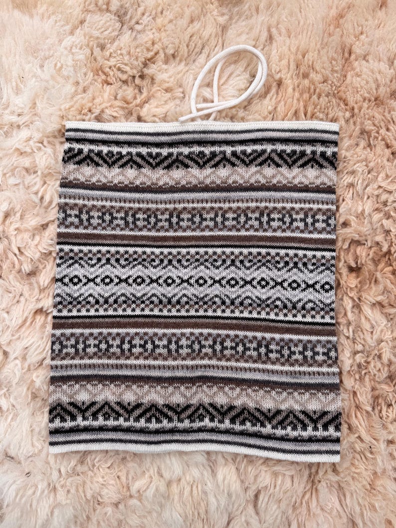 May include: A knitted neck gaiter with a geometric pattern in shades of brown, gray, and white. The gaiter has a white drawstring at the top. The background is a textured, light brown surface.