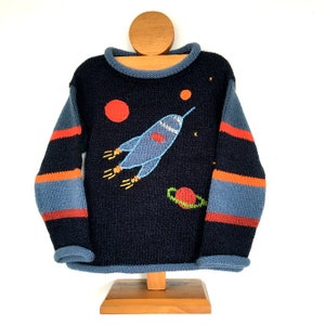 Könnte beinhalten: Ein marineblauer Strickpullover mit orangefarbenen und blauen Streifen an den Ärmeln. Der Pullover zeigt eine gestickte Rakete, Planeten und Sterne.