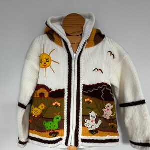 May include: White knitted hooded jacket with brown trim and embroidered animal and sun designs. The jacket features a zipper closure and a brown and white striped pattern on the sleeves.
