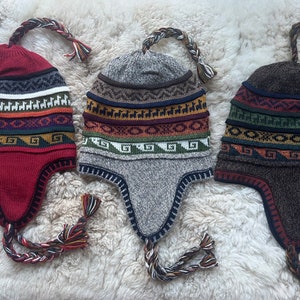 May include: Three colorful knitted winter hats with ear flaps and braided tassels. The hats feature a geometric pattern with stripes and a llama design.