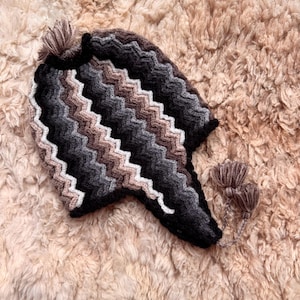 May include: A knitted hat with a zig-zag pattern in black, grey, white, and brown. The hat has ear flaps and a tassel on top, with two more tassels attached to the ear flaps. The hat is displayed on a textured, light brown surface.