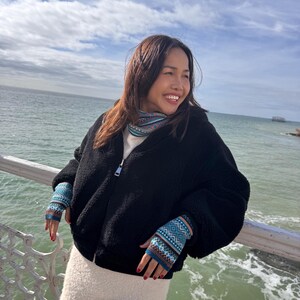 May include: A black fleece zip-up jacket with a white top and a white skirt. The person is wearing blue and teal patterned fingerless gloves and a patterned scarf. The background shows the ocean and a cloudy sky.