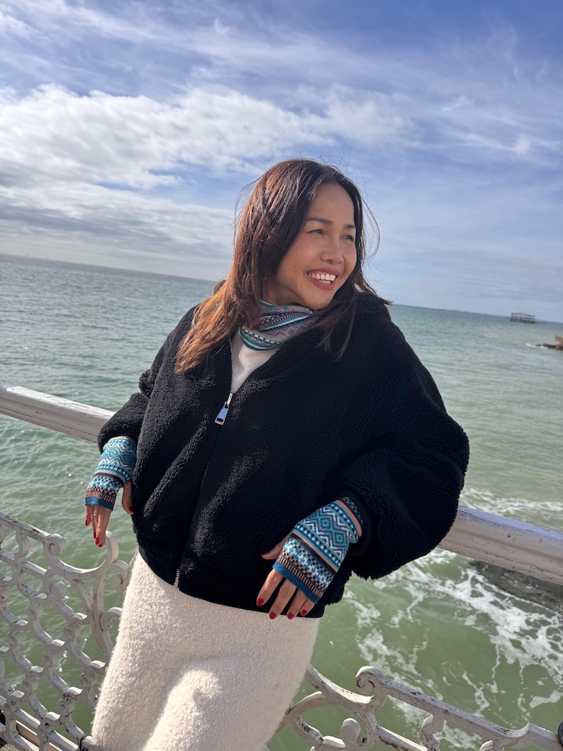 May include: A black fleece zip-up jacket with a white top and a white skirt. The person is wearing blue and teal patterned fingerless gloves and a patterned scarf. The background shows the ocean and a cloudy sky.