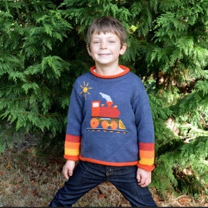 May include: A young boy wearing a blue knitted jumper with a red train design and orange and yellow stripes on the sleeves. He is wearing blue jeans and standing in front of a green bush.