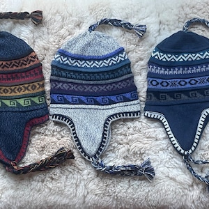 May include: Three knitted winter hats with ear flaps and braided tassels. The hats are in different colors and feature geometric patterns.